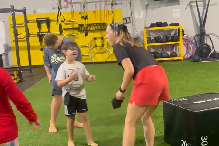 A coach in red shorts and a young boy in glasses grin at each other during a chase game on the turf