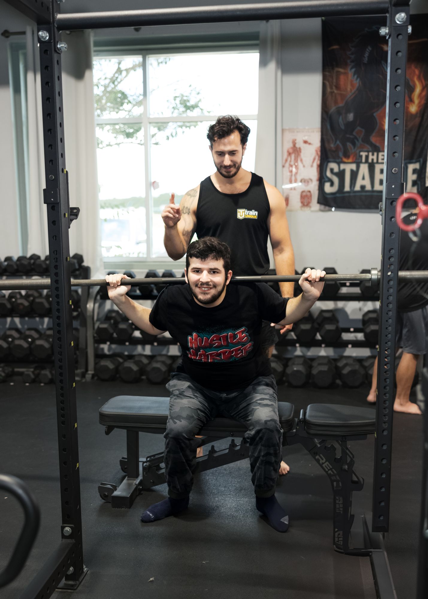 An athlete smiles from under the bar in a squat rack while his coach stands behind him, one finger raised