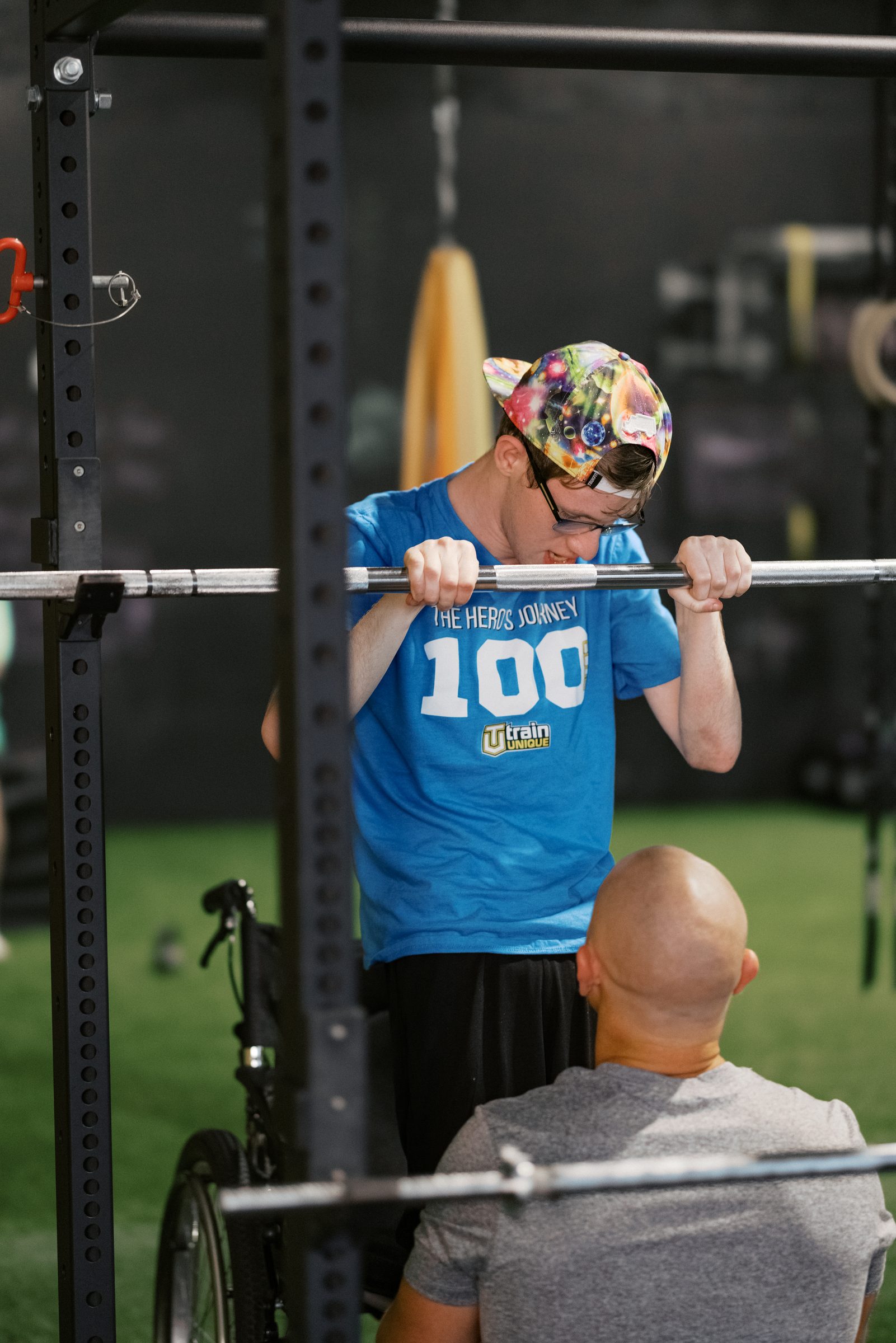 An athlete in a Hero's Journey 100 shirt grips the bar in the squat rack, his coach right in front of him