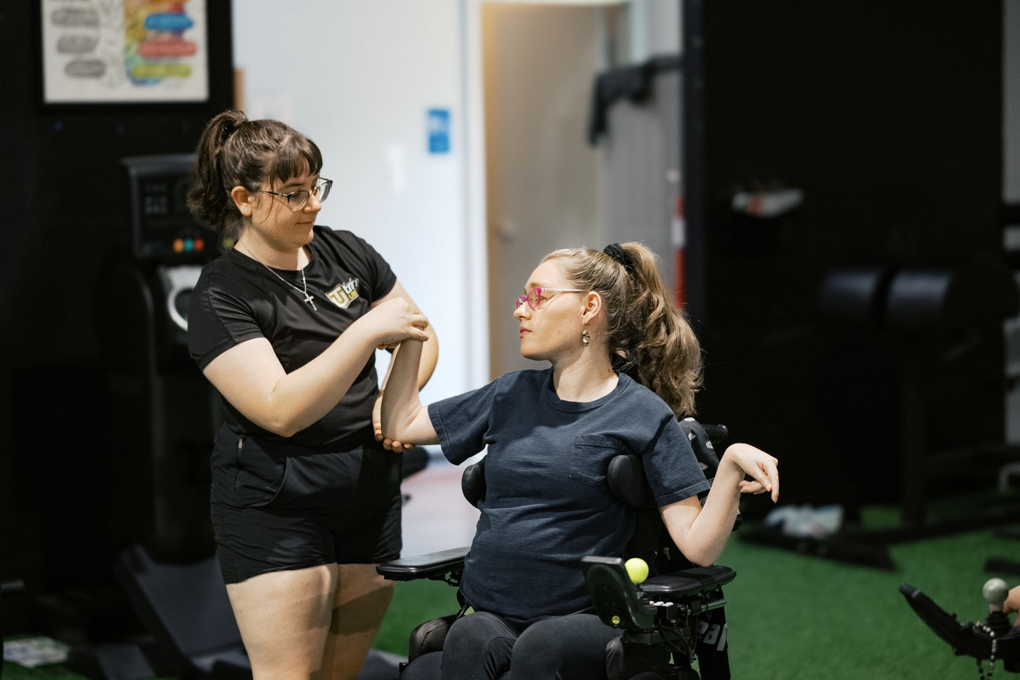A coach holds an athlete's elbow to guide an arm movement while the athlete, seated in a power wheelchair, looks at her